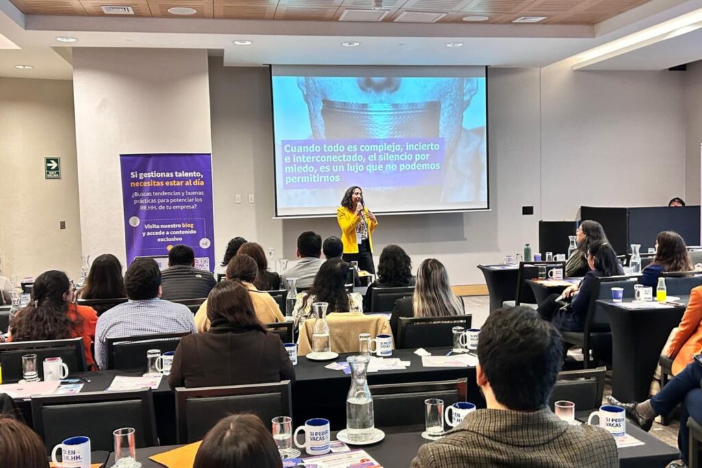 Fotografía de Paola Canessa impartiendo una conferencia sobre liderazgo y comunicación en entornos complejos, vistiendo saco amarillo, frente a una audiencia atenta en un salón corporativo.
