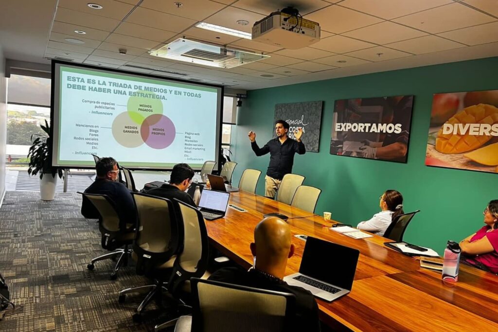 Fotografía de Patricio García impartiendo una capacitación empresarial sobre estrategias de medios en una sala de juntas moderna, con proyector y asistentes tomando notas.