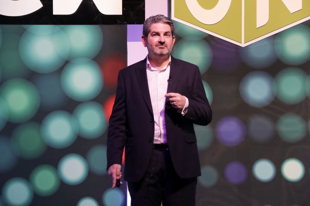 Fotografía de Pedro Eloy durante una conferencia, vistiendo traje formal oscuro y camisa clara, en un escenario con fondo colorido y luces desenfocadas que aportan dinamismo.