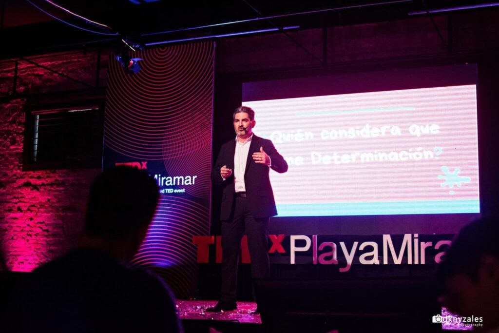 Fotografía de Pedro Eloy durante su ponencia en TEDx Playa Miramar, vistiendo traje formal oscuro y hablando con determinación en un escenario iluminado con luces púrpuras y rosas.