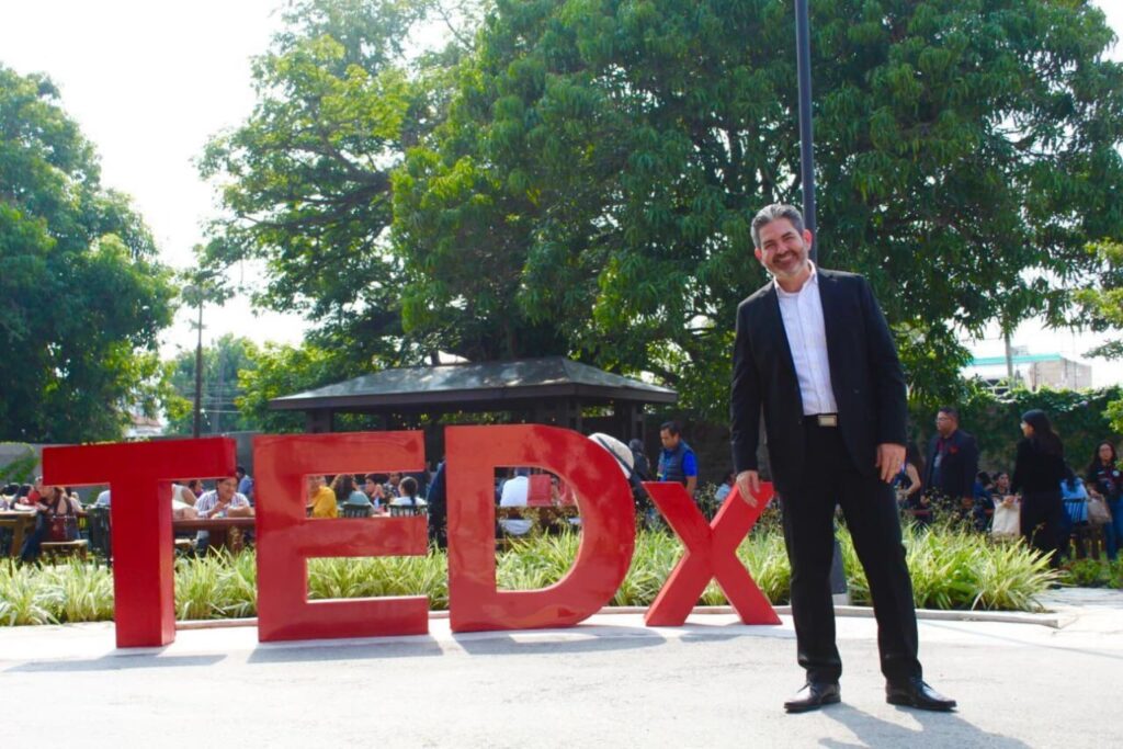 Fotografía de Pedro Eloy junto al icónico letrero rojo de TEDx en un evento al aire libre, con un ambiente de networking y asistentes en segundo plano.