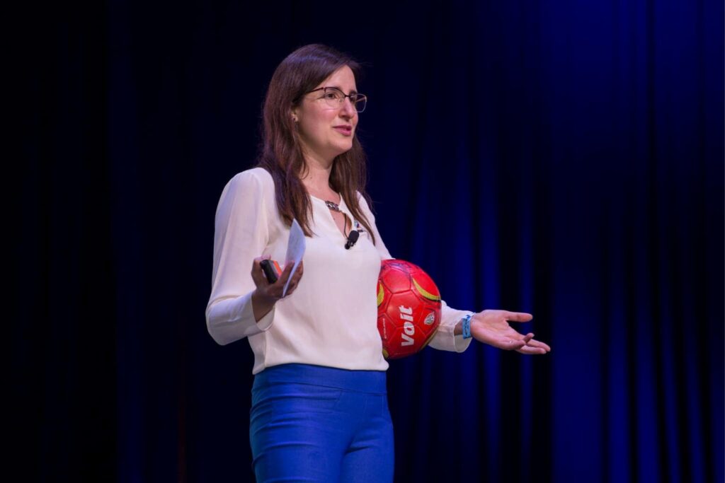 Fotografía de Ximena Hernández Buffa durante una conferencia, sosteniendo un balón de fútbol rojo mientras expone frente a un público en un escenario iluminado con fondo azul oscuro.