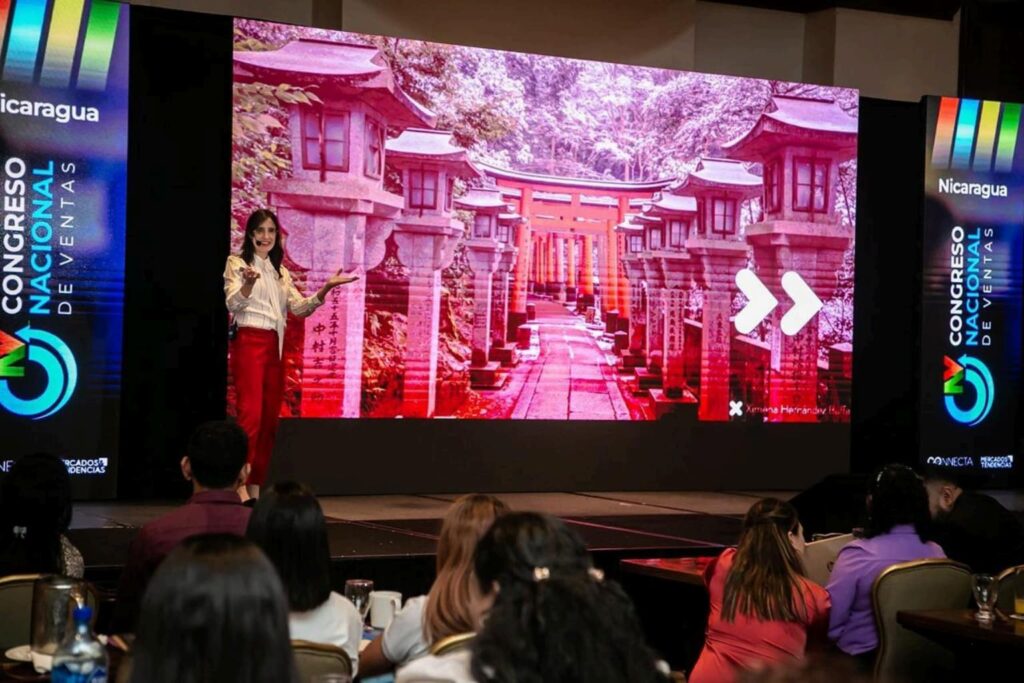 Fotografía de Ximena Hernández Buffa durante su ponencia en el Congreso Nacional de Ventas en Nicaragua, mostrando una diapositiva con un pasillo de templos orientales, mientras comparte estrategias de ventas ante un público atento.