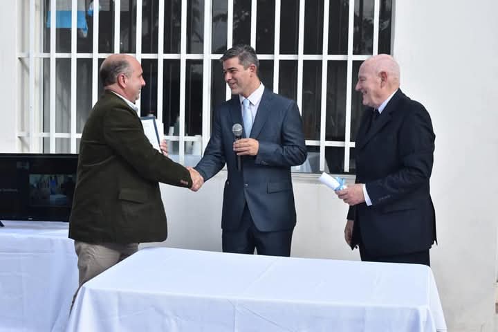 Lucas Romero Sosa en un acto institucional al aire libre, vestido con traje formal y micrófono en mano, estrechando la mano de un participante mientras otro hombre observa con una carpeta.