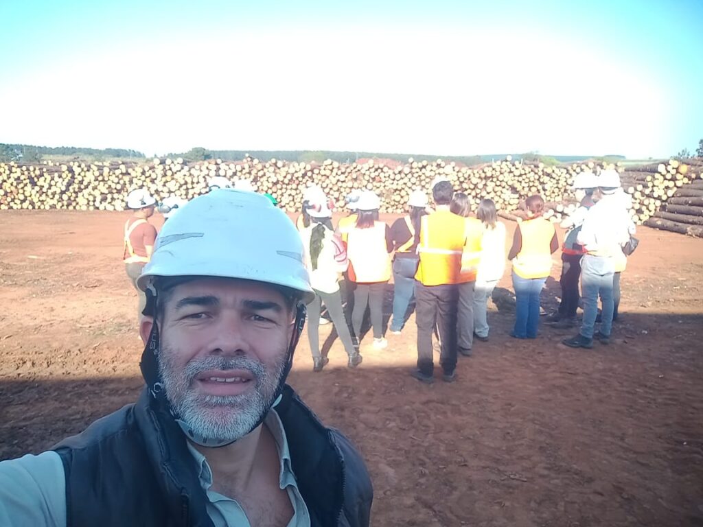 Lucas Romero Sosa en una planta forestal al aire libre, usando casco de seguridad y chaleco, con un grupo de personas al fondo en visita técnica frente a pilas de troncos apilados.