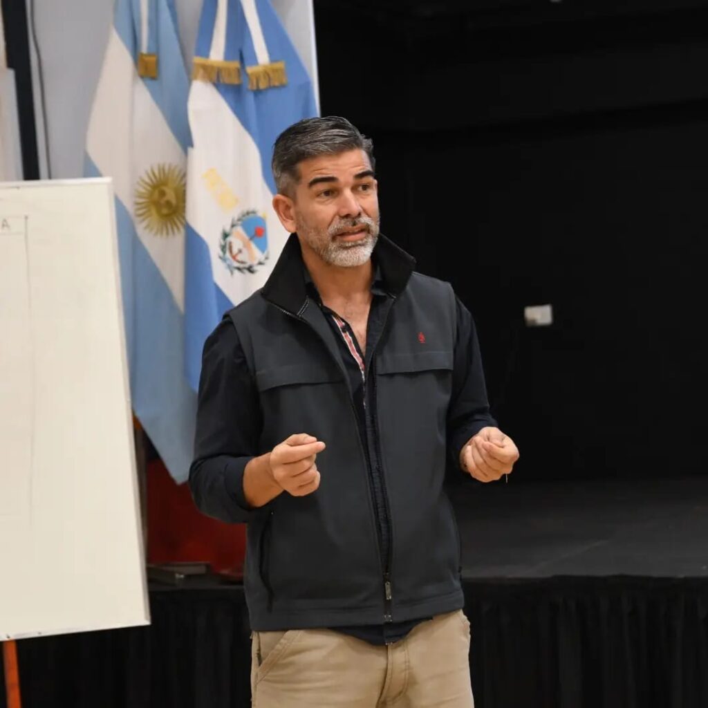 Lucas Romero Sosa dando una charla en un auditorio con banderas argentinas al fondo, vistiendo chaleco oscuro y gesticulando mientras se dirige al público.