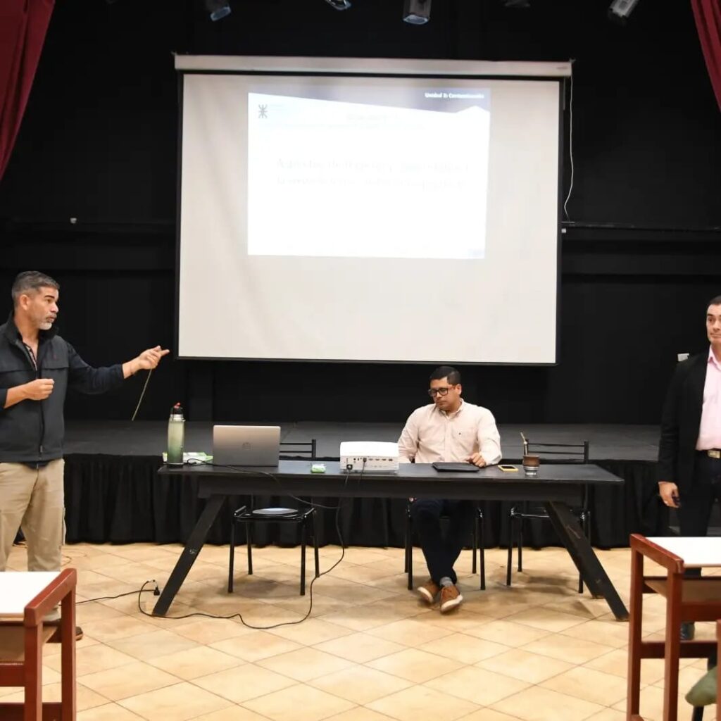 Lucas Romero Sosa durante una presentación académica en un auditorio, señalando hacia una pantalla de proyección junto a dos colegas sentados en una mesa con equipos tecnológicos.