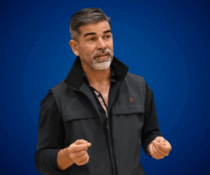 Lucas Romero Sosa durante una conferencia, con fondo azul sólido, luciendo chaleco oscuro y gesticulando con las manos en actitud expresiva y comunicativa.