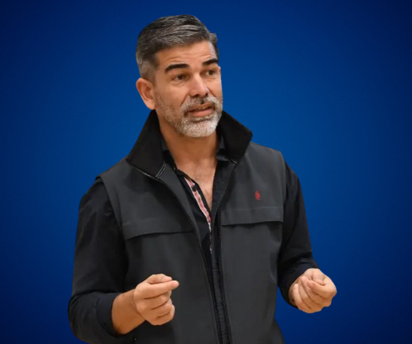 Lucas Romero Sosa durante una conferencia, con fondo azul sólido, luciendo chaleco oscuro y gesticulando con las manos en actitud expresiva y comunicativa.