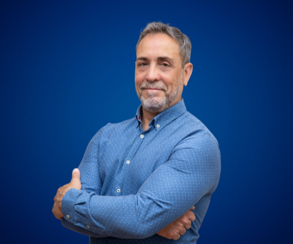 Marcelo Carbone, de pie con los brazos cruzados, vistiendo una camisa azul con fondo azul sólido. Su expresión transmite seguridad y profesionalismo.