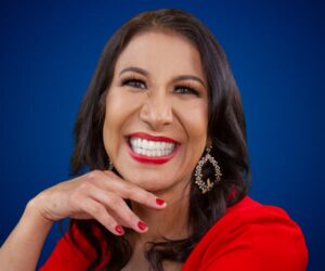 Fotografía de María Antonieta Alcalá con vestido rojo, sonriendo con expresión vibrante frente a un fondo azul, con aretes llamativos y manicura roja.