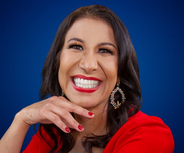 Fotografía de María Antonieta Alcalá con vestido rojo, sonriendo con expresión vibrante frente a un fondo azul, con aretes llamativos y manicura roja.