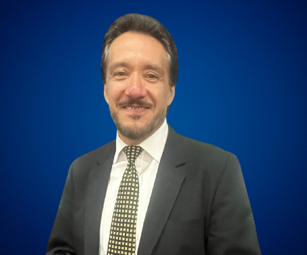 Fotografía de Ness Carballo, vistiendo traje formal con corbata de patrón geométrico, posando con una sonrisa sobre un fondo azul liso.