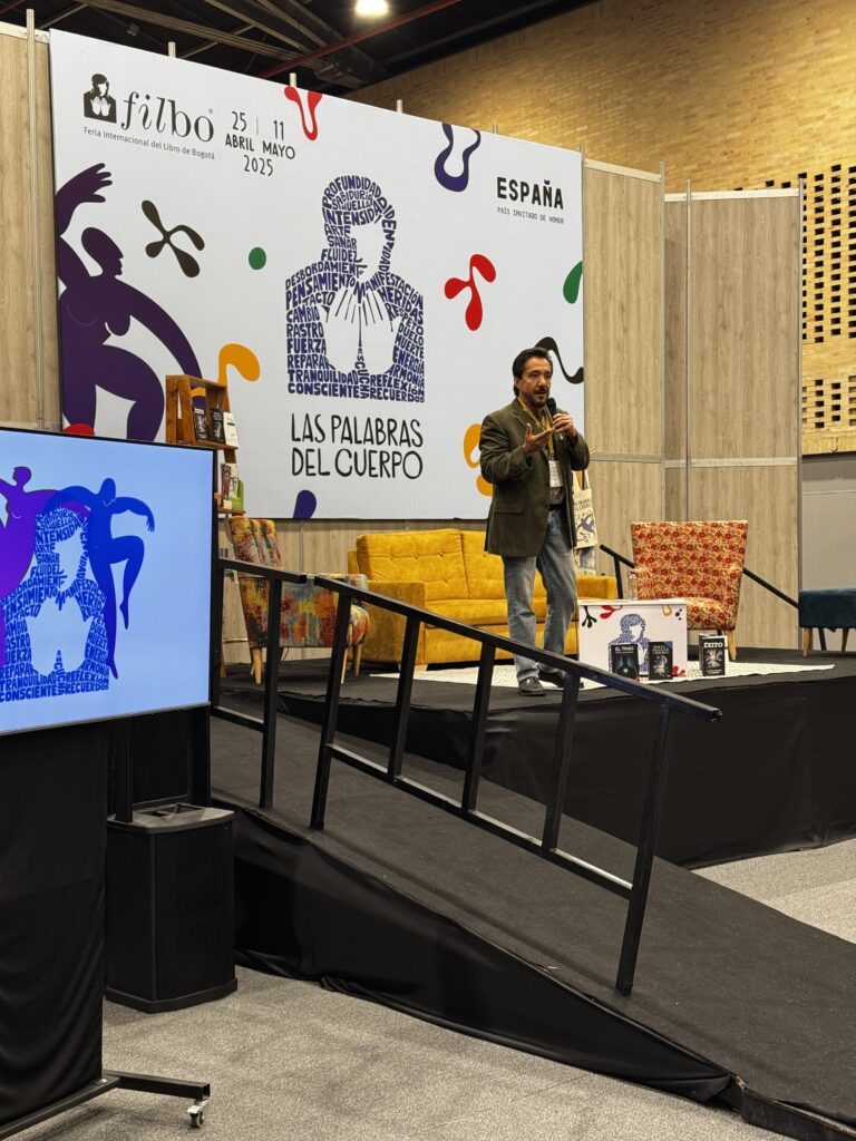Ness Carballo en la Feria Internacional del Libro de Bogotá 2025, de pie sobre un escenario con micrófono en mano, frente a un colorido fondo con la temática "Las palabras del cuerpo".