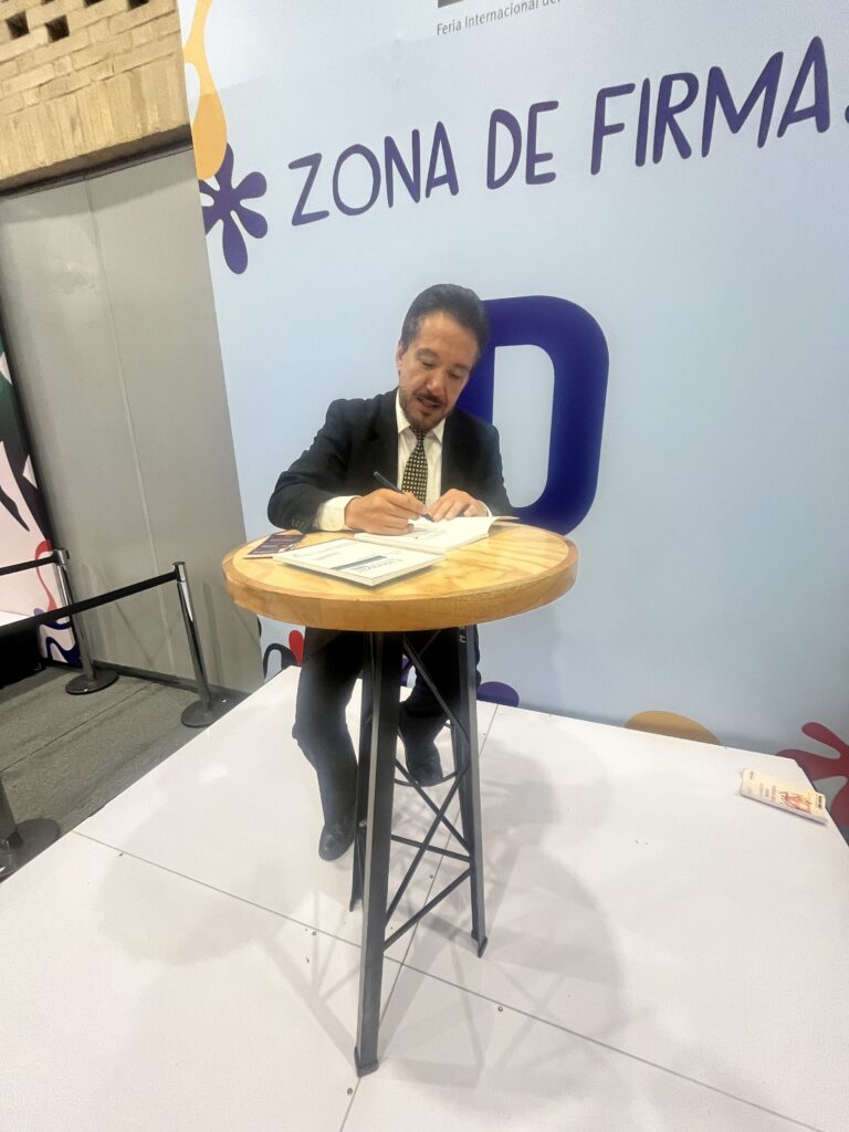 Ness Carballo en la zona de firmas de la Feria Internacional del Libro de Bogotá, escribiendo dedicatorias en libros sobre una mesa alta de madera.