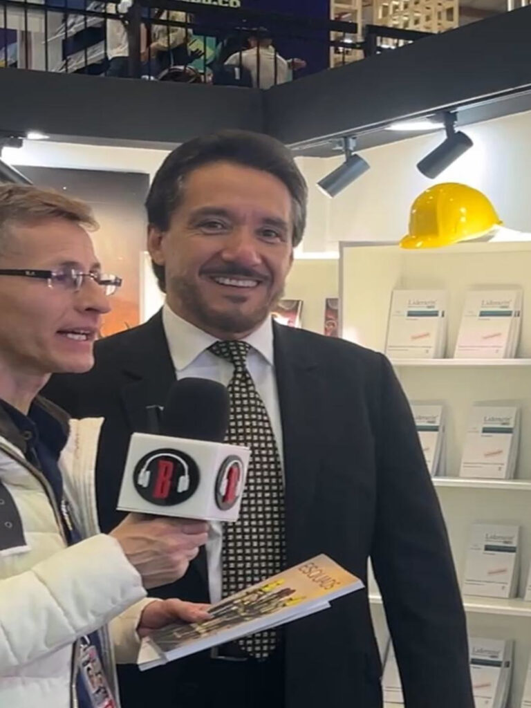 Fotografía de Ness Carballo durante una entrevista en un evento editorial, vistiendo traje formal y corbata, acompañado de un periodista con micrófono y una revista en mano. Al fondo, estantería con libros y un casco de seguridad amarillo como elemento decorativo.