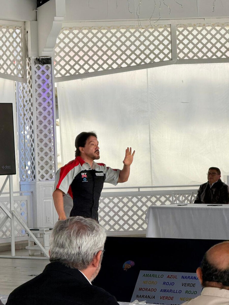 Fotografía de Ness Carballo impartiendo una conferencia en un espacio cerrado con estructura blanca y estilo colonial. Viste uniforme con logos institucionales mientras gesticula con la mano levantada frente a un público atento.