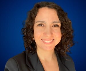 Fotografía de Paola Canessa con una sonrisa cálida, vistiendo un saco oscuro y posando sobre un fondo azul profesional.