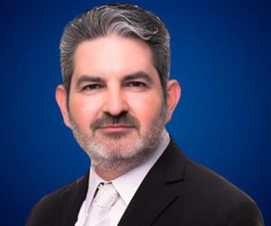 Fotografía de Pedro Eloy con fondo azul, vistiendo traje formal oscuro, camisa blanca y corbata clara, transmitiendo profesionalismo y confianza.