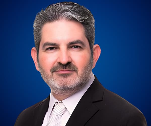 Fotografía de Pedro Eloy con fondo azul, vistiendo traje formal oscuro, camisa blanca y corbata clara, transmitiendo profesionalismo y confianza.