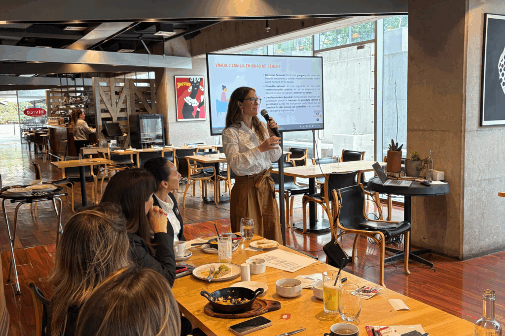 Fotografía de Carolina Pinto Grez dando una charla en un restaurante moderno, de pie frente a una pantalla con presentación sobre equidad de género, mientras varias personas la observan atentamente.