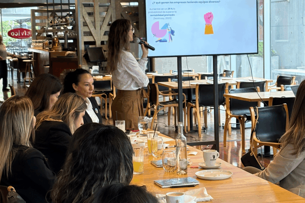 Fotografía de Carolina Pinto Grez presentando una diapositiva sobre diversidad en equipos, frente a un grupo de mujeres profesionales en un restaurante moderno y bien iluminado.