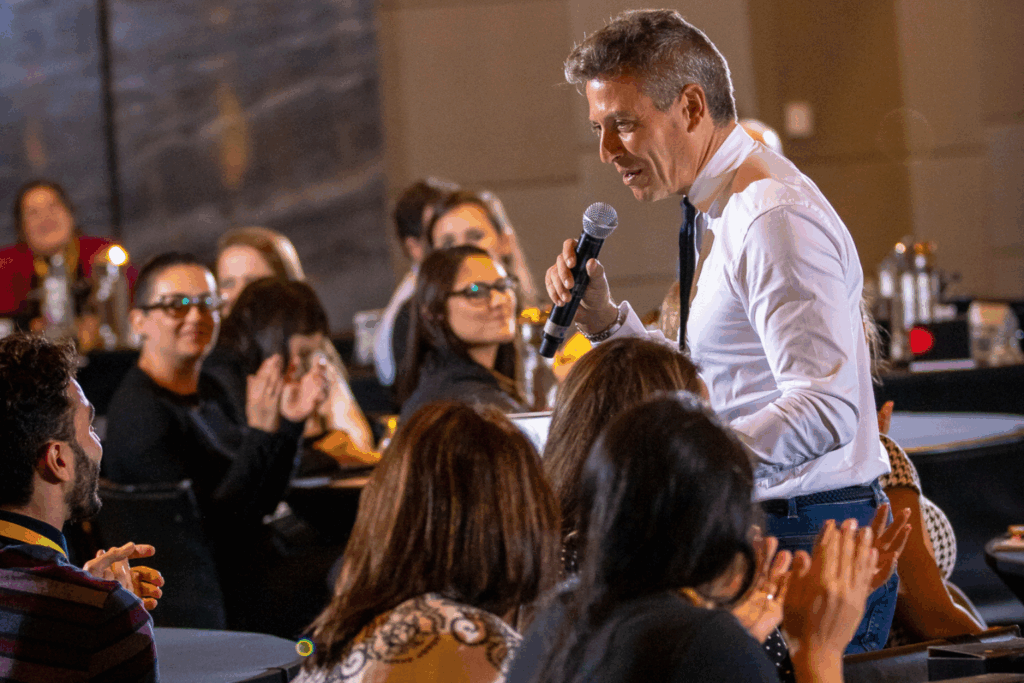 Fotografía de Enric Company durante una conferencia interactiva, sosteniendo un micrófono mientras se dirige al público con cercanía y entusiasmo. La audiencia lo observa con atención y aplaude su intervención.