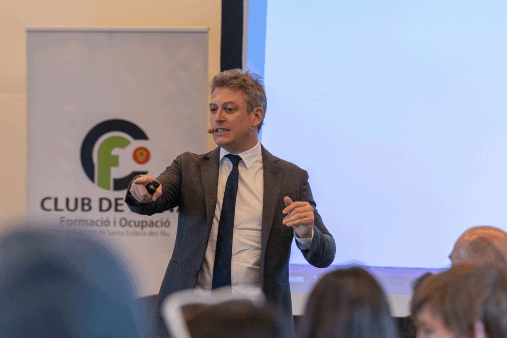 Fotografía de Enric Company dando una conferencia en un auditorio, señalando hacia el público con mando en mano, vestido formalmente frente a una pantalla y un banner institucional.