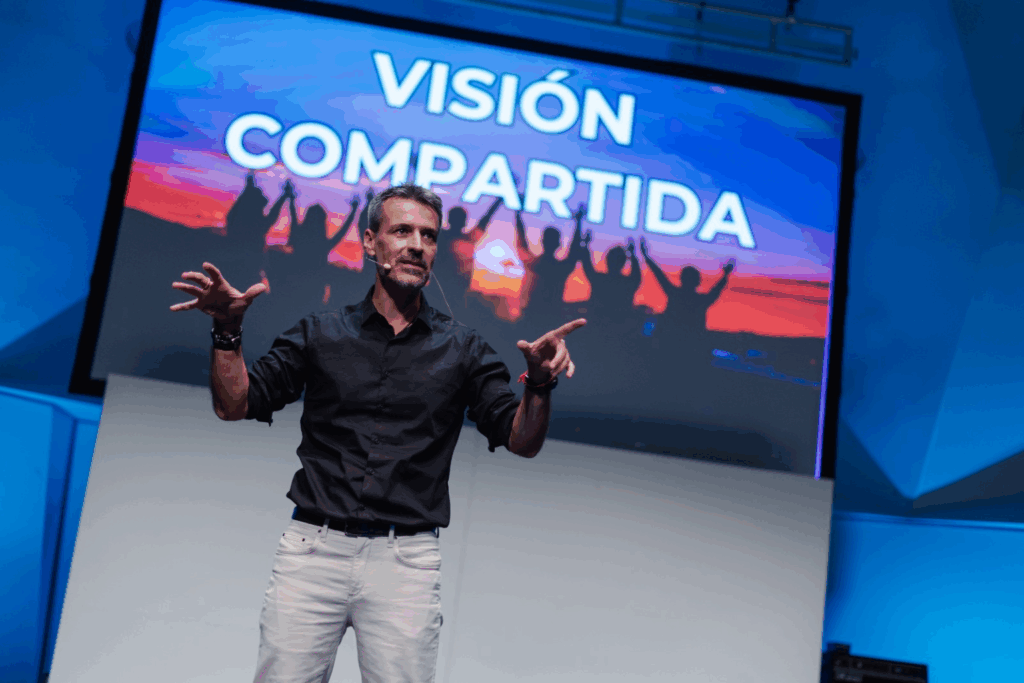 Fotografía de Antoni Fernández i Brugués en el escenario durante una conferencia, con fondo azul y una gran pantalla que muestra la frase "Visión Compartida" sobre una imagen de personas al atardecer.