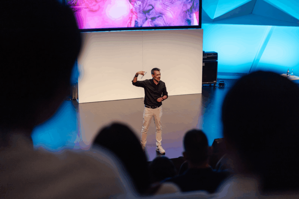 Fotografía de Antoni Fernández i Brugués de frente sobre el escenario, hablando con expresión enérgica ante una audiencia, con una pantalla al fondo que muestra una imagen emotiva de un niño y una persona mayor.