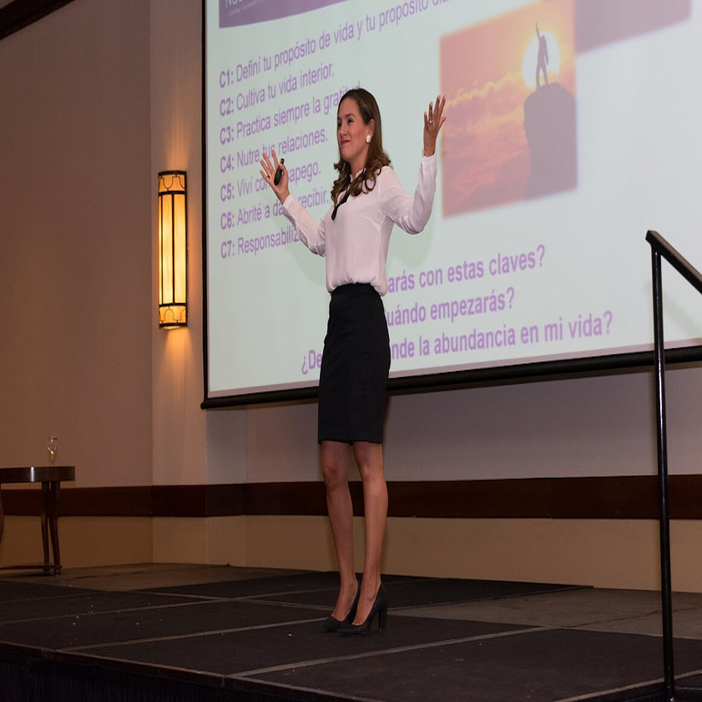Fotografía de Nadia Vado en una conferencia en interiores, de pie sobre un escenario, vestida con blusa blanca y falda oscura, con los brazos extendidos frente a una pantalla con texto motivacional.