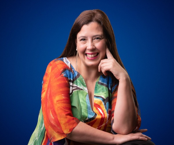 Fotografía de Emilse Silvestre, sonriendo con expresión cálida, vestida con una blusa colorida frente a un fondo azul sólido.