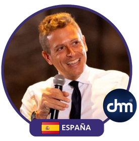 Fotografía de Enric Company durante una conferencia, sonriendo mientras sostiene un micrófono. Viste camisa blanca y corbata oscura, con fondo desenfocado y cálido, transmitiendo cercanía y entusiasmo.