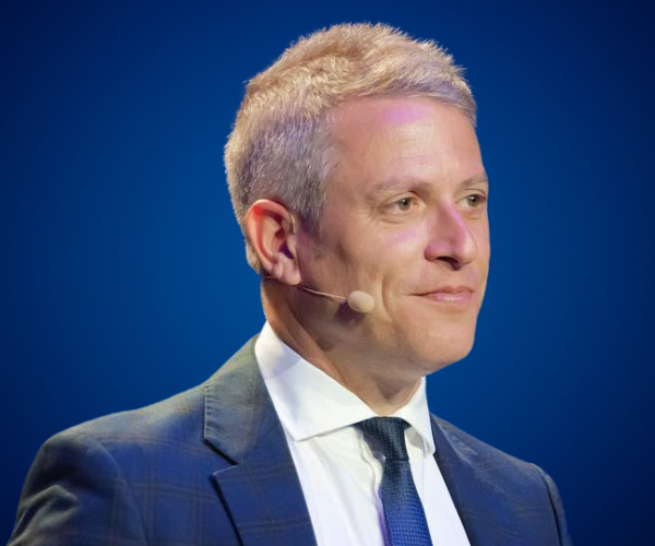 Fotografía de Enric Company en plano medio, luciendo traje formal y micrófono de diadema, sobre fondo azul intenso. Su expresión serena y confiada transmite profesionalismo y motivación.