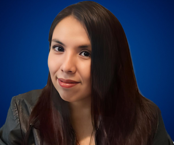 Fotografía de Erika Hernández Mejía con fondo azul, luciendo un abrigo verde oliva y una expresión segura, con la mano en el rostro en pose reflexiva.