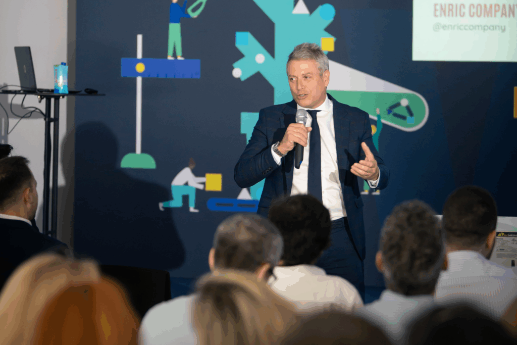 Fotografía de Enric Company durante una conferencia profesional, hablando con micrófono frente a una audiencia atenta. Viste traje formal y se encuentra frente a un fondo moderno con ilustraciones corporativas.