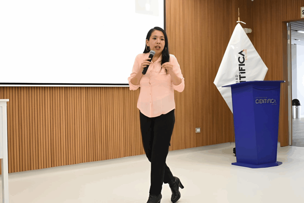 Fotografía de Erika Hernández, conferencista, hablando con micrófono en mano en un auditorio moderno de madera clara y fondo institucional.