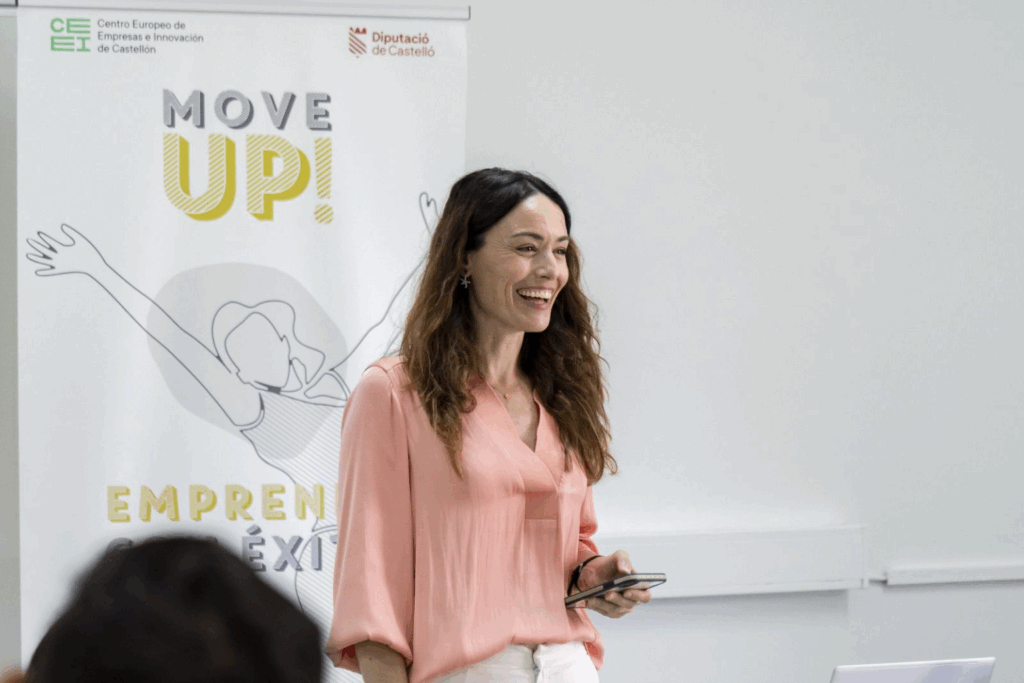 Fotografía de Eva Mazuecos durante una ponencia en el programa "Move Up!" del CEEI Castellón, sonriendo con una actitud cercana y positiva, vestida con blusa rosa y pantalón blanco, sosteniendo un móvil frente a un cartel institucional.