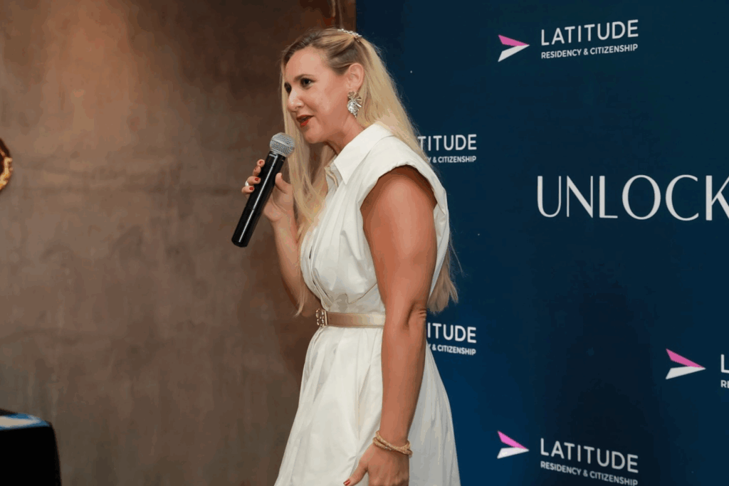 Fotografía de Meli Alemán durante una conferencia, vestida con un elegante conjunto blanco y hablando con micrófono en mano frente a un panel de fondo corporativo.