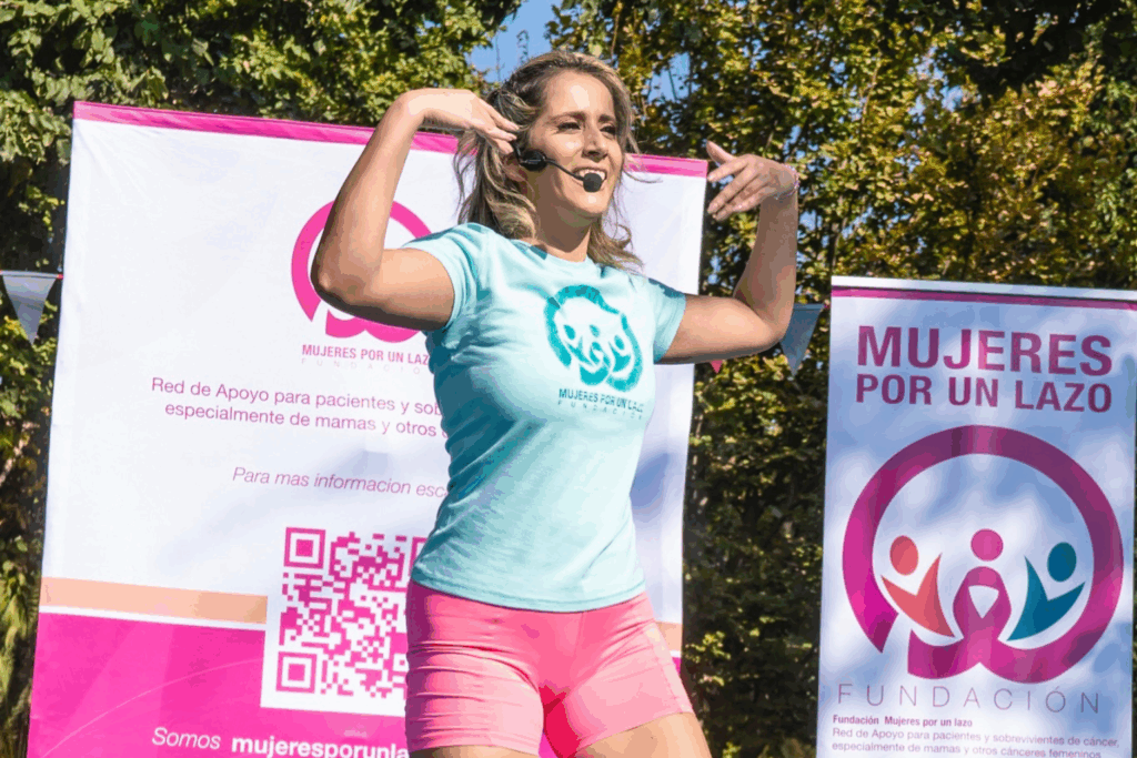 Fotografía de Mila Correa liderando una actividad al aire libre con micrófono de diadema, frente a banners de la Fundación Mujeres por un Lazo, con árboles de fondo.