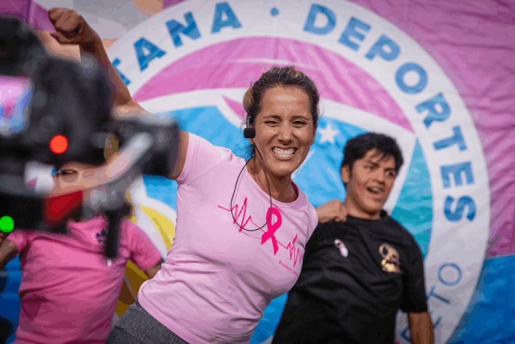 Fotografía de Mila Correa liderando una clase de actividad física con temática de concientización sobre el cáncer de mama, vestida con camiseta rosa y micrófono, en un evento deportivo comunitario.