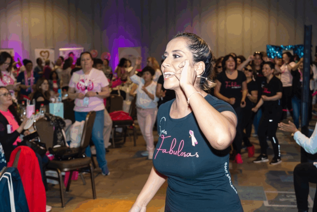 Fotografía de Mila Correa guiando una dinámica motivacional con mujeres en un evento cerrado, todas con atuendos alusivos a la lucha contra el cáncer de mama.