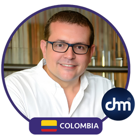 Fotografía de Gustavo De La Vega, conferencista colombiano, con camisa blanca y gafas, sonriendo en un entorno interior iluminado con fondo moderno de madera y cristal.