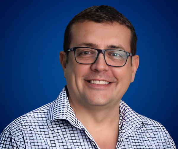 Fotografía de Gustavo De La Vega con camisa a cuadros blancos y negros, gafas de montura oscura y fondo azul profesional