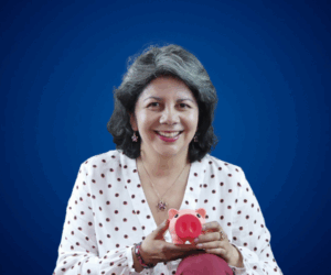 Fotografía de Lyda Ardila sosteniendo una alcancía rosada en forma de cerdito, vestida con blusa blanca de lunares y fondo azul, con una expresión cálida y profesional.