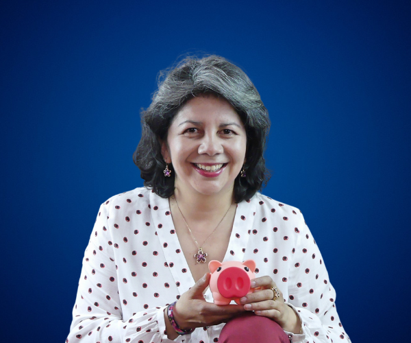 Fotografía de Lyda Ardila sosteniendo una alcancía rosada en forma de cerdito, vestida con blusa blanca de lunares y fondo azul, con una expresión cálida y profesional.