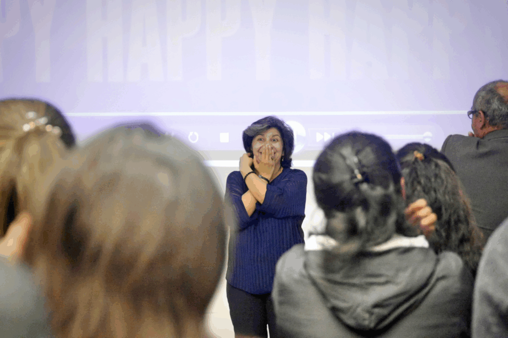 Fotografía de Lyda Ardila en plena dinámica con un grupo de asistentes, mostrando un gesto expresivo de entusiasmo frente a una pantalla con la palabra “HAPPY”.