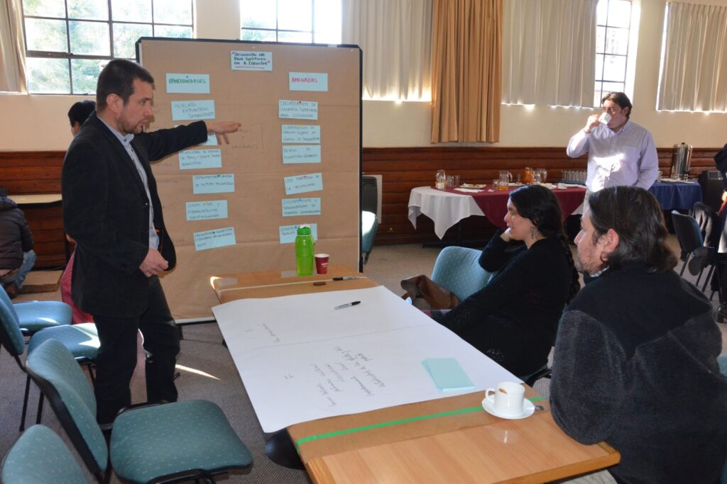 Fotografía de Mauricio Espina dirigiendo un taller en sala de reuniones, señalando un panel con notas organizadas mientras interactúa con participantes sentados alrededor de una mesa de trabajo.