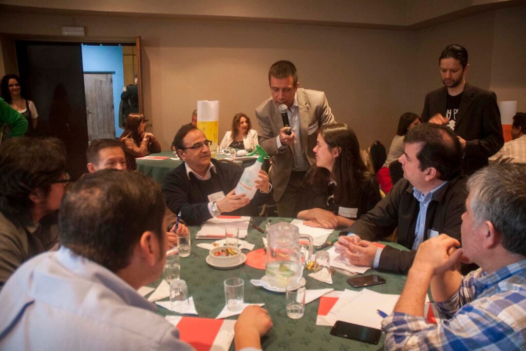 Fotografía de Mauricio Espina facilitando un taller grupal, con micrófono en mano, interactuando con participantes que presentan un producto en una mesa de trabajo colaborativa.