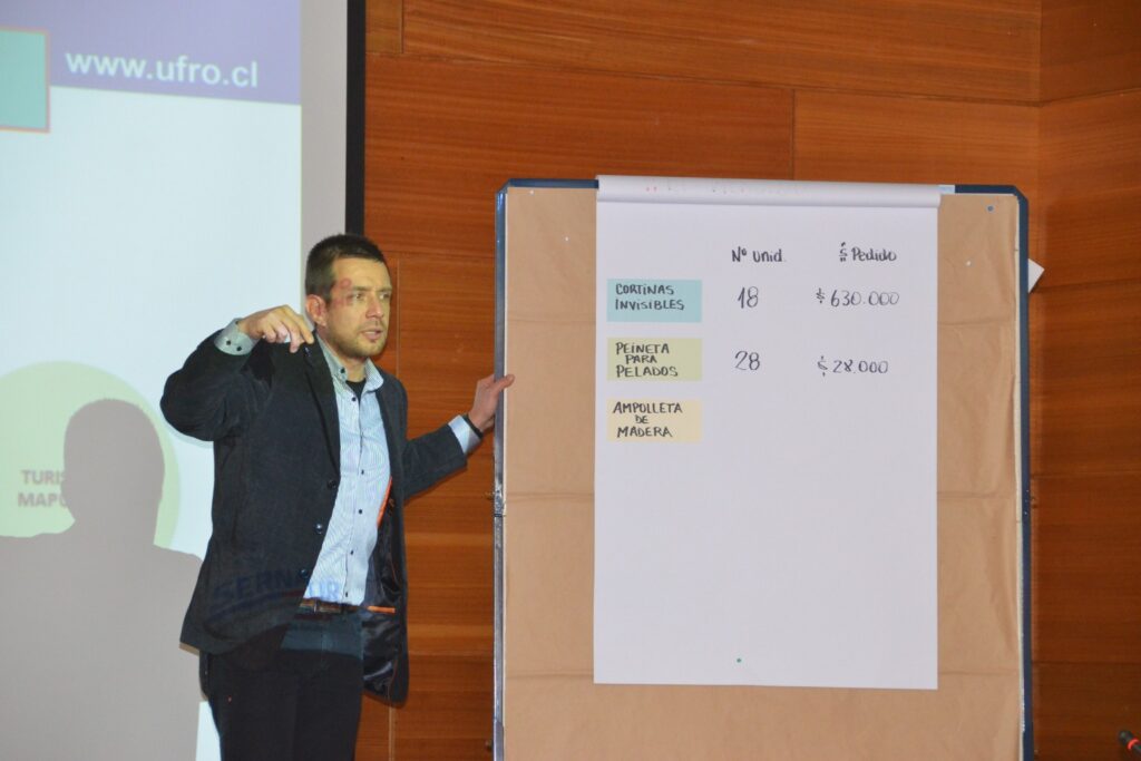 Fotografía de Mauricio Espina en sala de conferencias, de pie junto a una pizarra con ejercicios escritos, explicando conceptos frente a un público en un entorno académico y profesional.